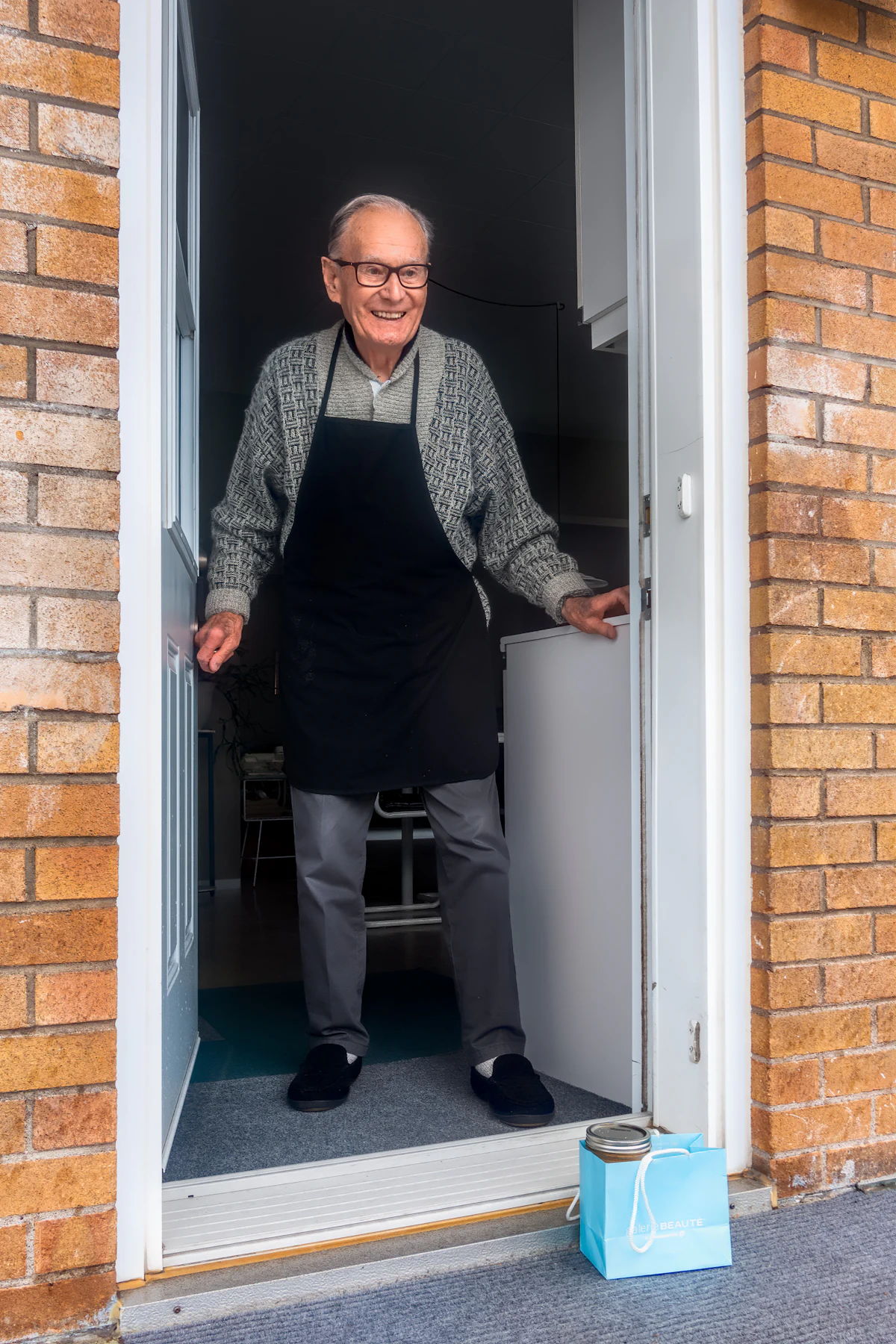An older man in an apron standing in the doorway of his shop